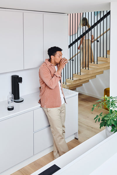 A man drinking sparkling water in a white kitchen with a black Mysoda Toby sparkling water maker standing on the kitchen counter